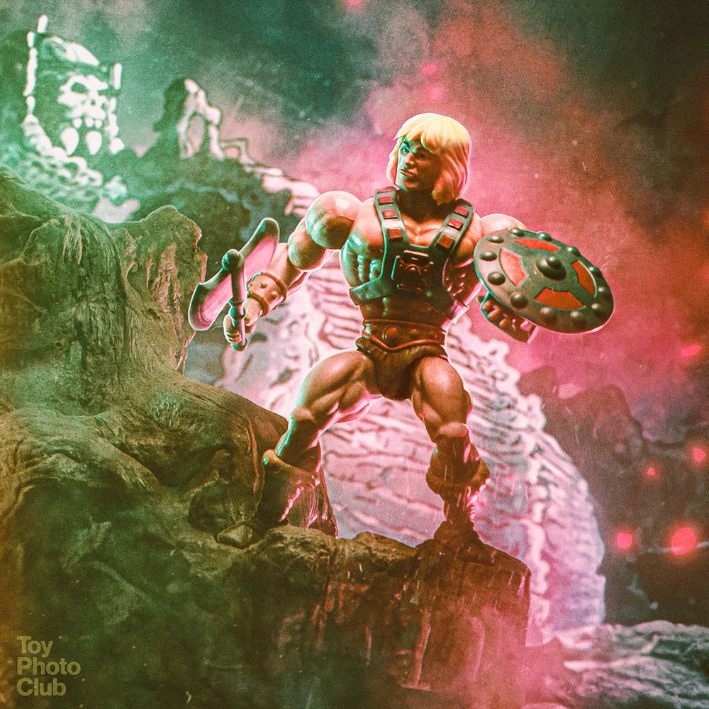 A toy photo of He-Man from Masters of the Universe. He's standing with a sword and shield on a rocky outcropping in front of Castle Grayskull.