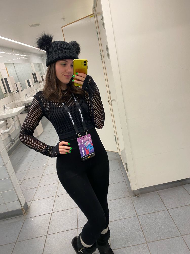 Yas wears black trousers, boots and strappy top, with fishnet long sleeves, and bobble hat. She is wearing a Dreamhack pink creator badge. She has shoulder length brunette hair and bright green nails. 