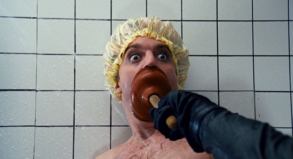 Beef, wearing a yellow shower cap and shocked expression, gets his mouth plunged by Winslow, the Phantom of The Paradise, while taking a shower in a white tiled bathroom. 