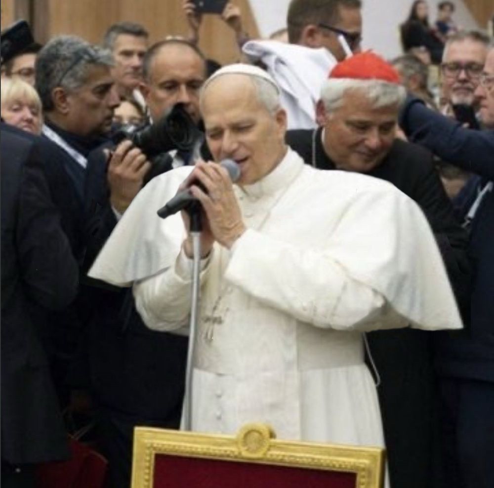 A photo of Pope Leo XIV on the mic surrounded by fellow God-bods and journalists. His shoulder pads are now comically oversized, in homage to David Byrne's Noh-inspired Talking Heads tour jacket.