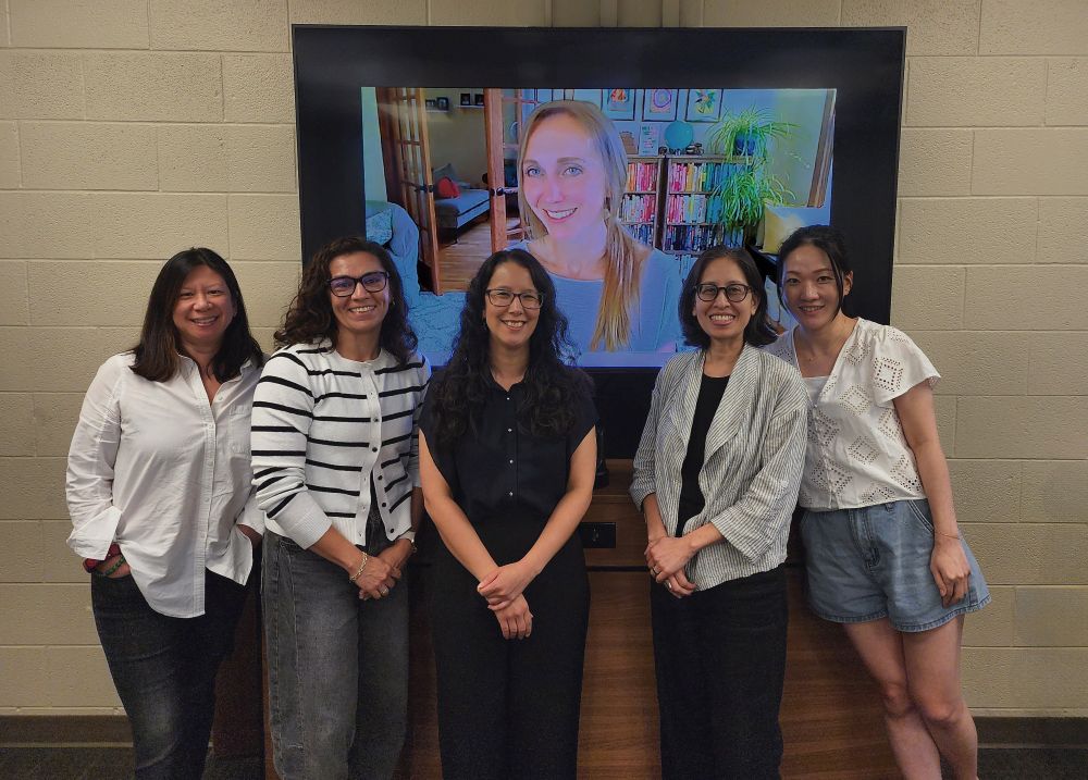 Group of 4 women smiling at the camera in front of a screen with a fifth woman on video call