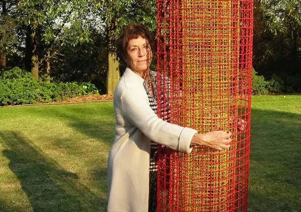 Helena Escobedo Hugs a piece of her art, as she gazes out at the viewer. They are in a garden with shrubs and trees beyond.