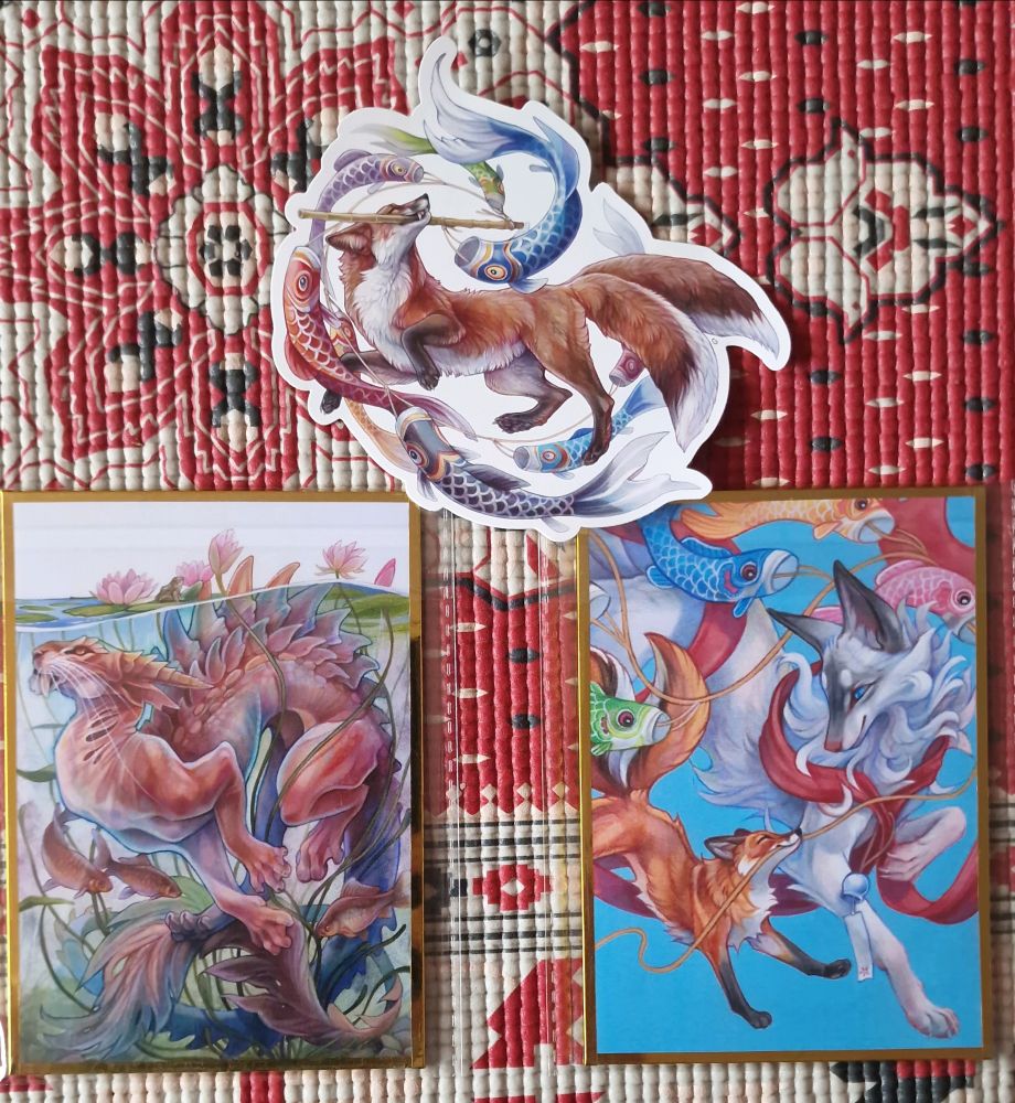 Photo of a large sticker and two gold-bordered mini prints by SushiArtStudio, one print shows a sabre-toothed feline draconic creature coiled beneath the surface of a pond surrounded by fish and water lilies, while the other print and the sticker are different illustrations of a red fox pulling the strings of carp-shaped kites, accompanied by a much larger white fox in the print.