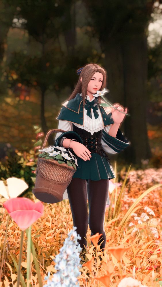 Camilla, wearing a white blouse with green accents, a matching green cape, and a black corset, puts a white flower up to her face as she walks in a forest meadow. She has a wicker basket of white flowers on her arm.