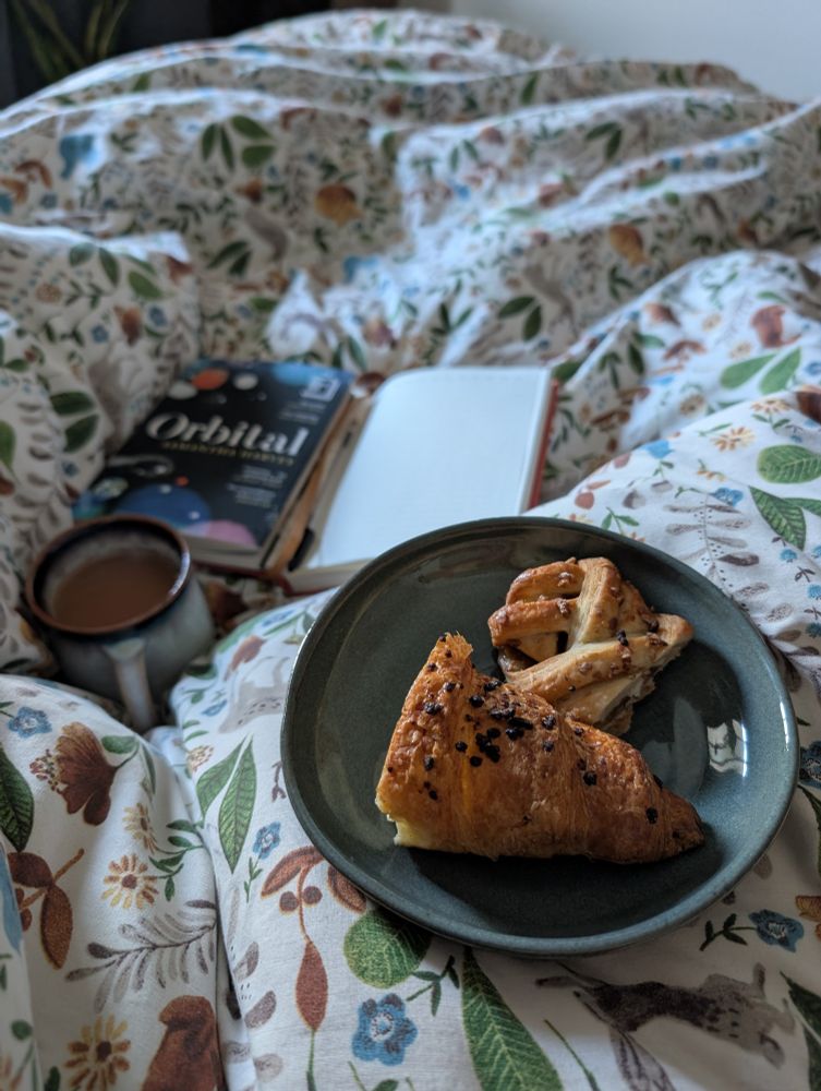 Breakfast in bed. Pastries on a plate in foreground, a mug of tea and a book in the background