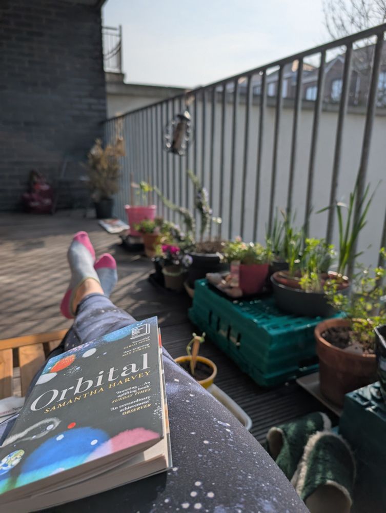 Sun soaked balcony, potted plants along railings. Foreground has a paper back copy of Orbital on top of legs stretched out 