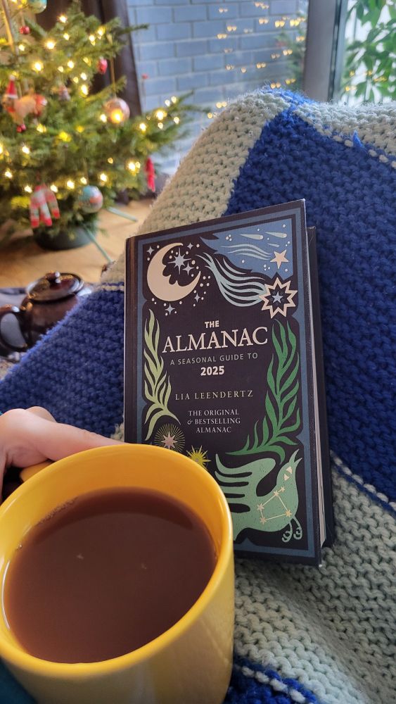 A large yellow mug of tea, the cover of 'The Almanac, A Seasonal Guide to 2025' both on a dark and light blue patchwork women blanket.  In the background, a brown teapot in front of a Christmas tree, lit up with fairy lights. 