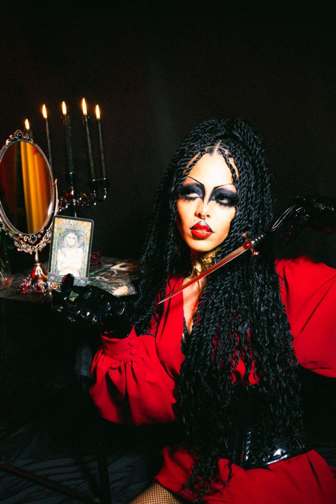Gothic black woman poses and holds a knife and a tarot card in front of a table with candles and a mirror. 