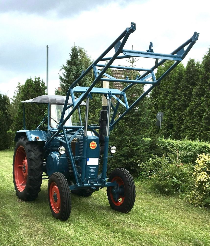 Das Foto zeigt einen blauen Lanz Bulldog D2806 mit roten Felgen, auf Rasen, vor Tannen und einer Hecke.