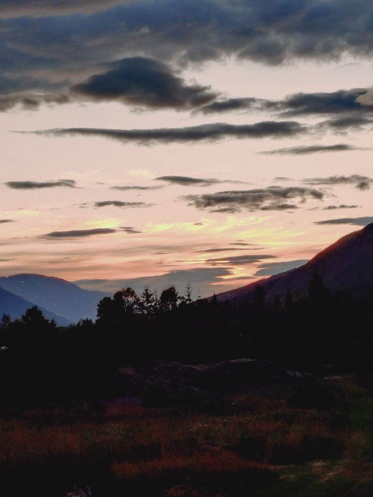 Dark landscape in the foreground, blue mountains in the distance. Above is the evening sky, pink and golden with a few small, dark gray clouds.
