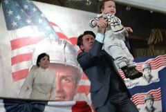 politician character in Dead Zone is wearing a dark colored suit on a campaign stage and holding up a toddler in front of him to dissuade a shooter from killing him. Mother of child is in background, wearing white, alarmed.  