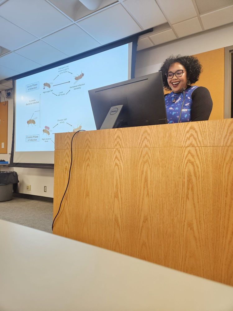 Dr. Darby (wearing a purple and blue fly dress with short black hair and square glasses) smiling behind a podium while presenting her dissertation.