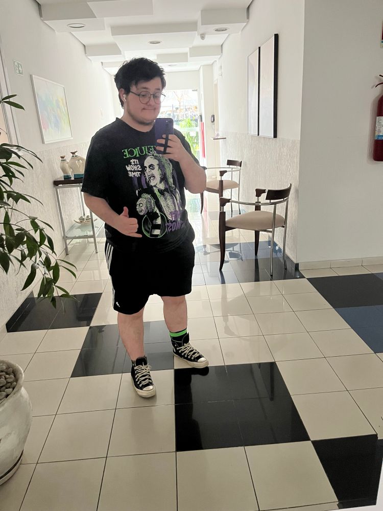 Estou em um hall de entrada de um prédio tirando foto no espelho. Sou branco com cabelo curto castanho e uso óculos. Estou vestindo uma camiseta preta com estampa do personagem beetlejuice, um short na altura do joelho preto e tênis all star de cano médio preto. Em uma mão seguro o celular pra foto e a outra faço sinal de hang loose.