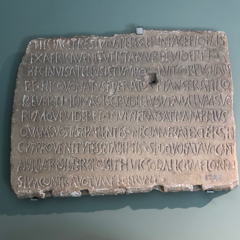 HIC IACET RESTUTUS PELEGER
IN PACE FIDELIS EX AFRICA VENIT
UT ISTAM URBE(M) VIDERET
H(A)EC INVISA TELLUS ISTUM VOLUIT CORPUS HABERE HIC
QUO NATUS FUERAT OPTANS
ERAT ILLO REVERTI ID MAGIS
CRUDELIUS UT NULLUM
SUORUMQUE VIDERET INVENERAT
SATIS AMPLIUS QUAM SUOS IPSE PARENTES NEC IAM ERAT EXTER
SICUT PROVENIT UT ESSET
AB IPSIS SED QUO FATA VOCANT
NULLUS RESISTERE POSSIT
HUICSODALICIUM FLORENSIUM
CONTRA VOTUM FECERUNT

"Here lies in peace the foreigner
Restutus, baptized. He came from Africa to see this city. But this unlucky land wanted his body.
From here he had wished to return to place of birth; it was all the more cruel, since he could not see any of his own. However, here he found much more than his parents; he was no longer a stranger as he had come, but considered one of us.
But when fate calls nobody can refuse to go. The Florenses association did this in spite of his hopes."

