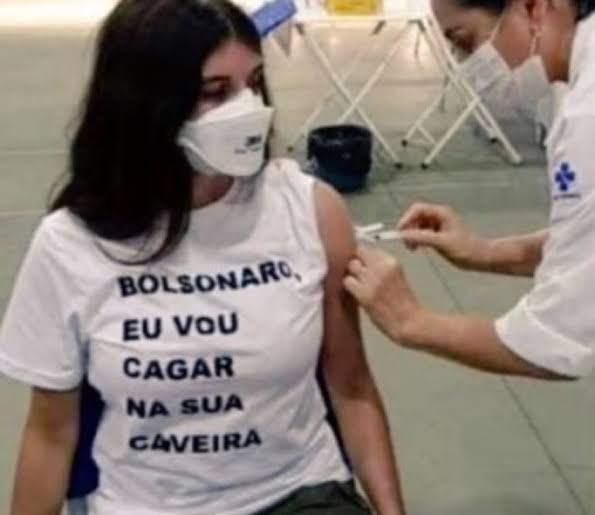 foto famosa da época da pandemia, uma garota com máscara respiratória toma vacina no SUS com uma camiseta escrito: "Bolsonaro eu vou cagar na sua caveira"