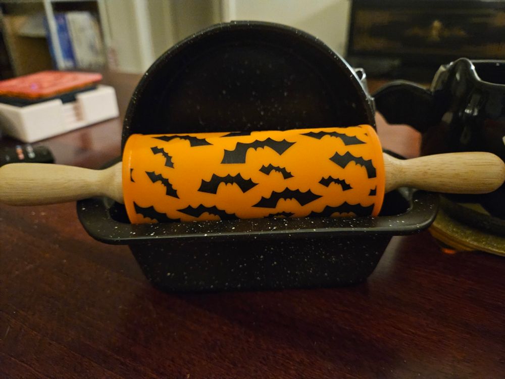 A set of miniature baking tools. There is a loaf pan, a round spring-release cake pan, and a rolling pin. The rolling pin has am orange silicone center part that has bats printed all over.