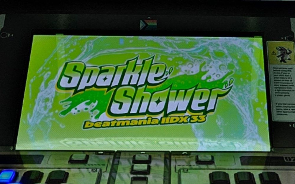 A pride sticker placed in the center above a Beatmania 2DX screen. My dancing donkey has been on the cabinet for about 3 years as part of the warning placard, while the pride and trans stickers get removed and replaced over and over.