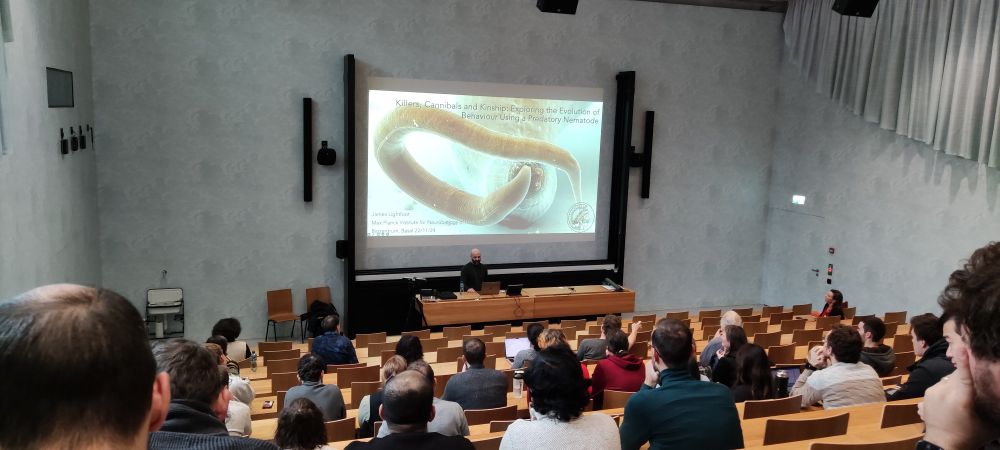 A picture of a lecture room full of people. There's a projected slide with a worm on it, and James is standing in front of it