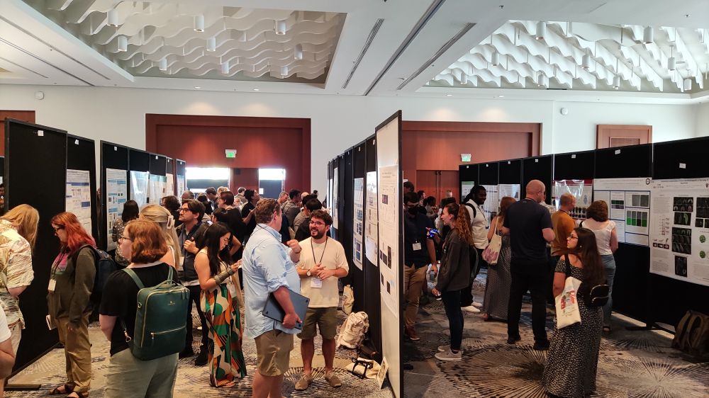 A picture of a poster session in a room. The session is full of people talking to each other and interacting in front of posters