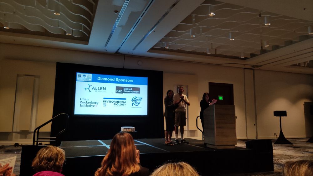 A picture of a projected slide with sponsors. Three people stand in front of the projected slide