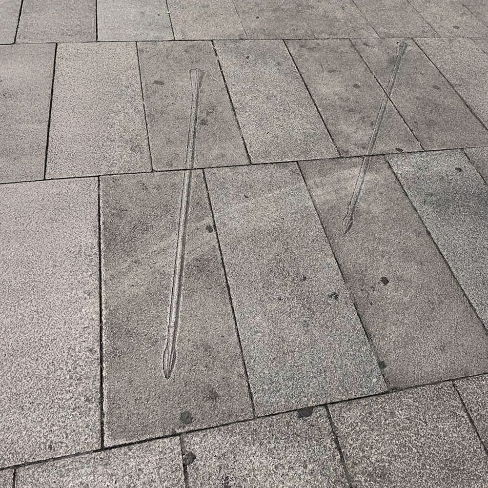 Imagen de un tramo del pavimento urbano de la Gran Vía madrileña, compuesto por grandes losas rectangulares de piedra gris. En dos de ellas se aprecian grabados lineales que representan flechas estilizadas, con punta afilada y plumas en el extremo. Las flechas están orientadas en direcciones ligeramente diferentes, como si hubieran sido disparadas y hubieran quedado clavadas simbólicamente en el suelo.