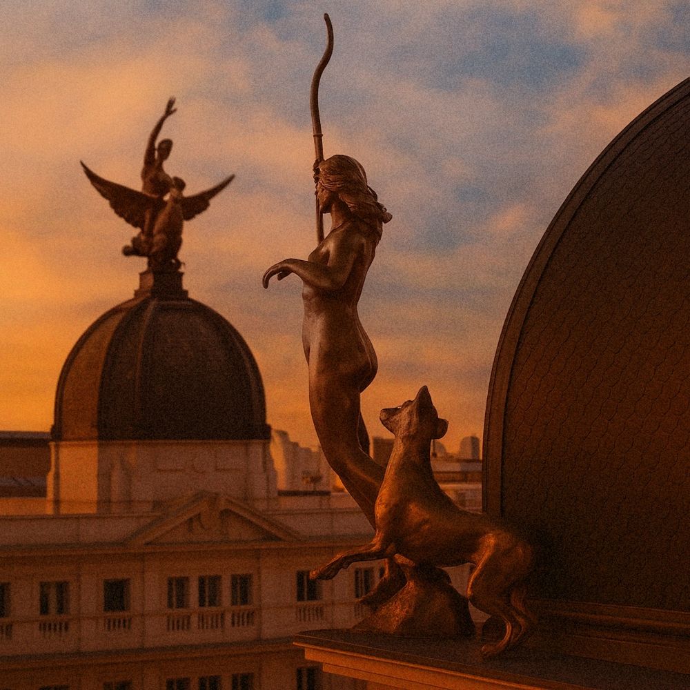 Escultura de Diana cazadora con un perro y un arco, al atardecer en una azotea de la Gran Vía de Madrid, apuntando hacia una figura alada sobre una cúpula del edificio opuesto.