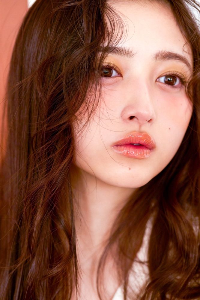 Nagi looking off into the distance thoughtfully, her bangs styled curly in the sides of her face, really glowy coral lip gloss on her lips