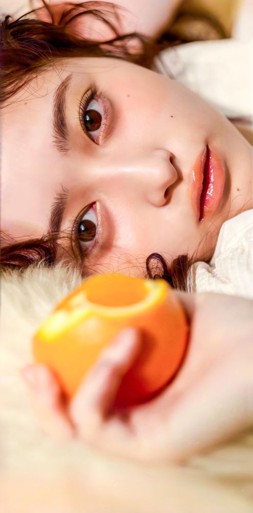 Closeup of Nagi’s face, lying horizontally in a fluffy rug or blanket, hand holding a partly chewed orange out of focus in the foreground