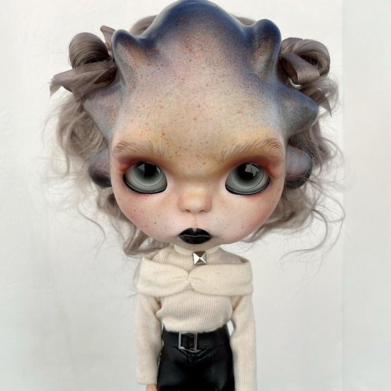 Custom blythe with heavily sculpted face. Protruding forehead with what looks like a bony casque. Stubby nose. Expression vaguely feels bulldog-like. Gray eyes with black eyeliner on the bottom. Short gray curly hair peaking from behind the casque.