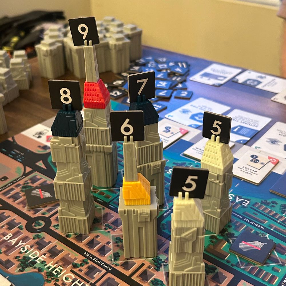 A game of High Rise in progress, with the UltraPlastic buildings. Stackable plastic buildings with a futuristic look on a board showing city streets. Each building has a top in an individual player color, and a chipboard square showing its height. 

On the board, a street named “Beela Boulevard” is visible. It’s named for one of our most beloved ferrets. She was incredibly empathetic and always seemed to know when we needed a bit of an emotional lift. 