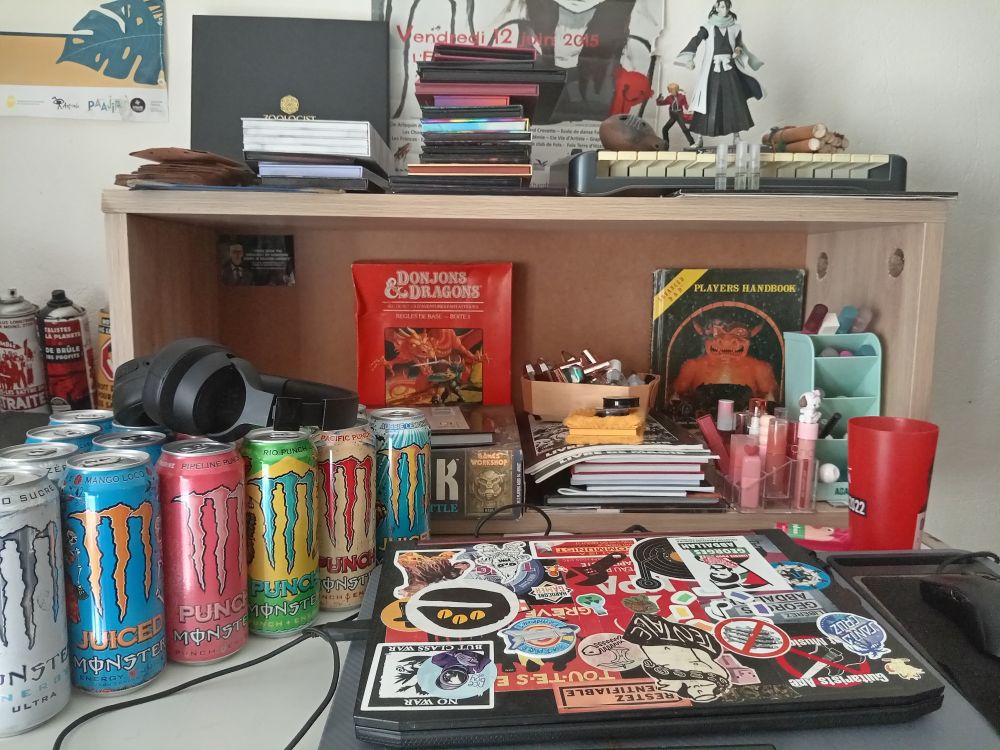 A gaming table with monster cans, old d&d books, cosmetics and a laptop