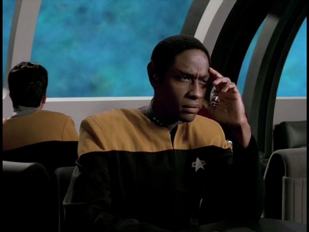 Tuvok sits at a table with one elbow resting on the table, his fingers pressed to his temple. He looks extremely tired and exasperated. Outside the window behind him, instead of the usual space, a black field with stars, there is a bluish greenish cloud. From an episode of Star Trek Voyager