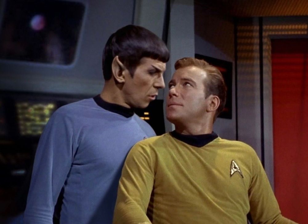 A very clever photo edit, made by some mad scientist trekkie, of Spock and Kirk from Star Trek TOS on the bridge of the Enterprise. Kirk is sitting in his Captain's chair, and Spock is standing directly behind him so that his chest is touching the back of Kirk's shoulder. Kirk's head is turned back towards Spock and Spock is leaning down over him as they gaze lovingly into each other's eyes. Their lips are only a couple inches apart, but they look like they are being drawn together by some magnetic force, into a kiss. 