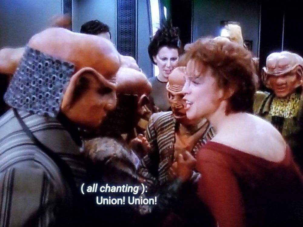 Screenshot from Star Trek DS9. Leeta, Rom, and the rest of the workers at Quark's Bar are all huddled together. Caption: 