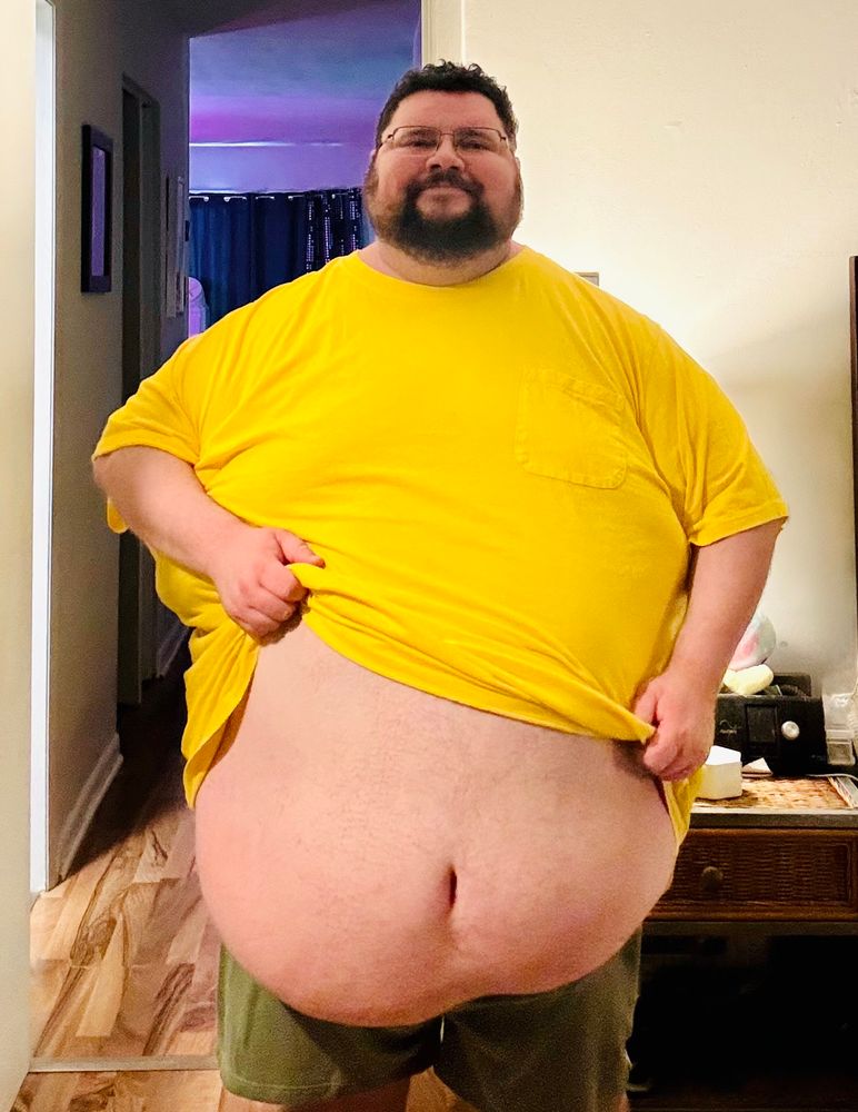 A very round and chubby scruffy guy wearing a yellow shirt is lifting his shirt to expose his very large fat belly. He looks pleased with himself. 