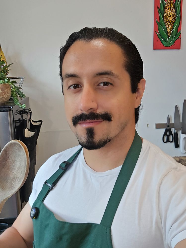 Twitch streamer with a green apron in a kitchen with kitchen equipment behind him house plants and a corn art, holding a wooden spoon.He has brown eyes, a mustache and long hair pulled back into a bun