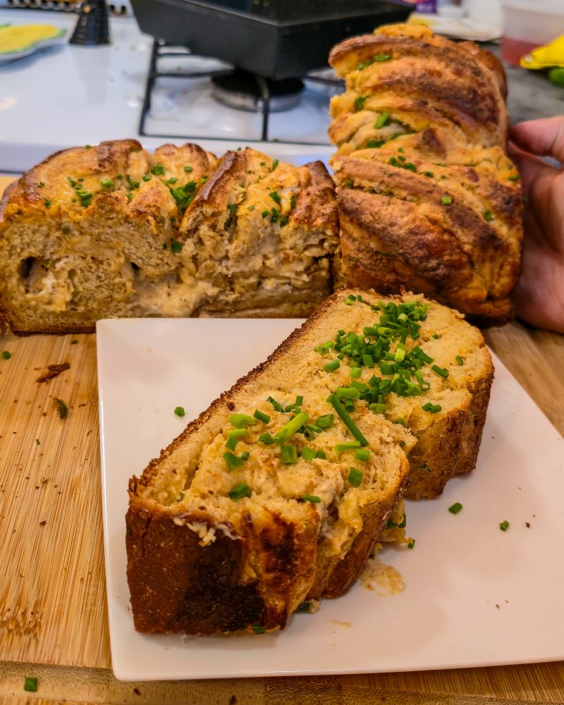 A golden brown and yellow blue bread that is stuffed with a crab dip with chopped chives, twisted and braided it on a white plate and wood cutting board freshly baked