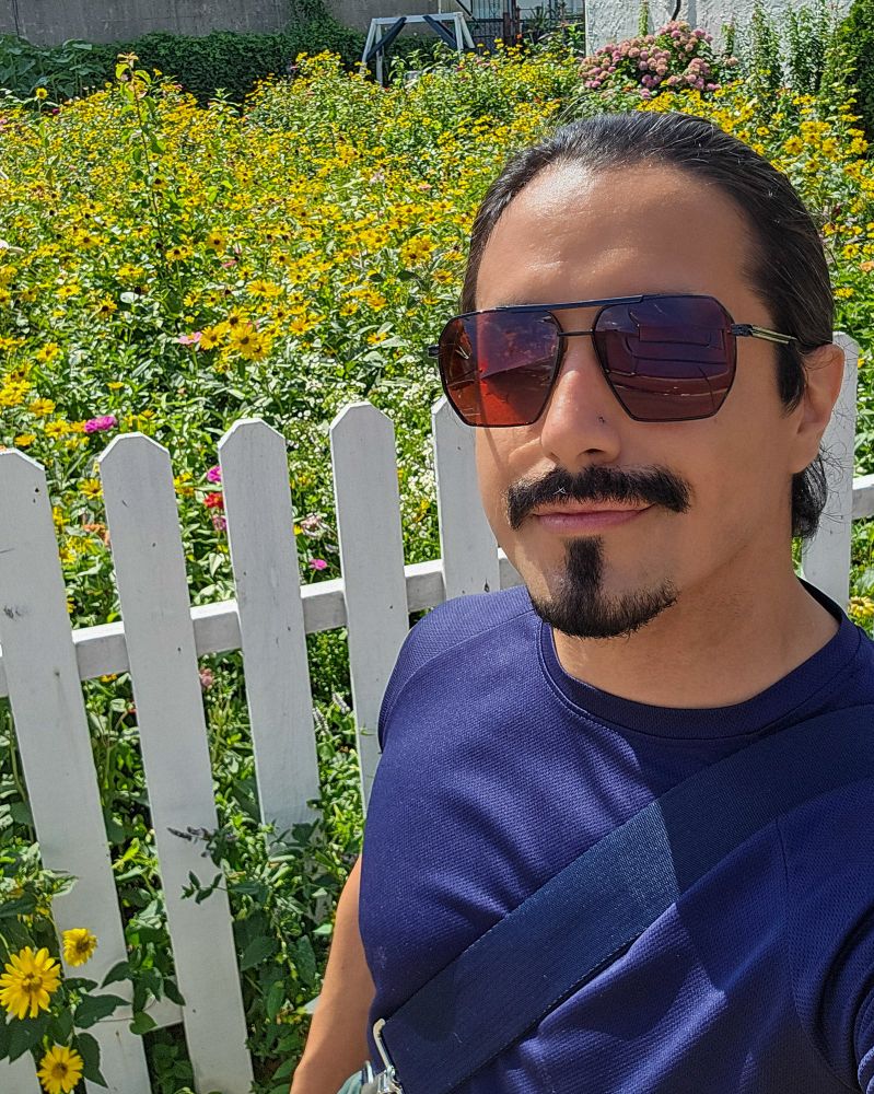 A man with a goatee and mustache and sunglasses with long black pulled back hair is standing in daylight in front of a lot of wildflowers with a white picket fence