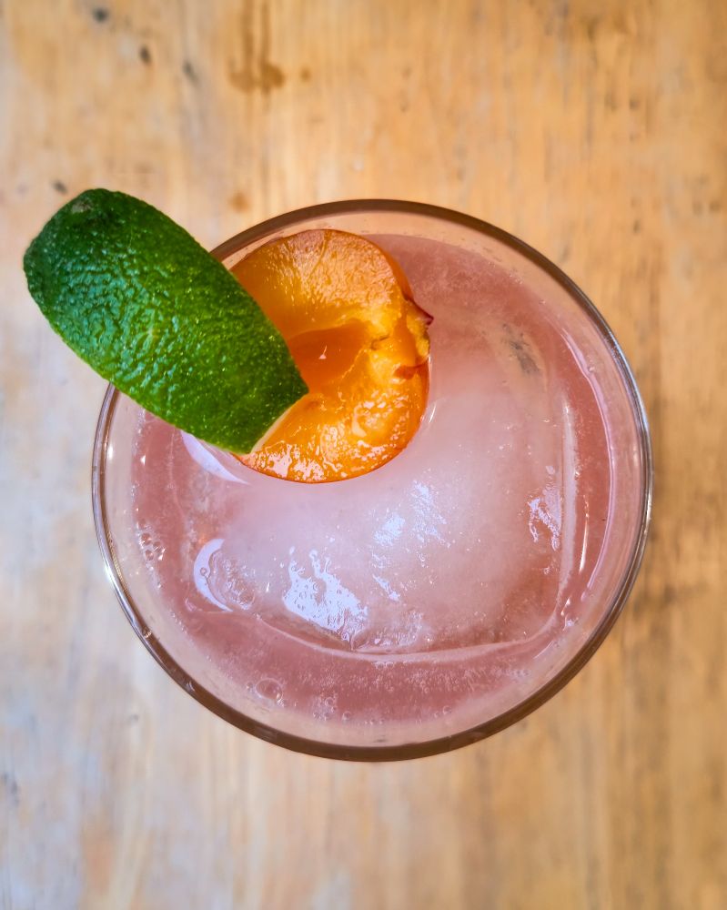 Overhead shot of a cocktail, but it's a golden plum. Soda with a large block of ice a half of a golden plum and a lime wedge