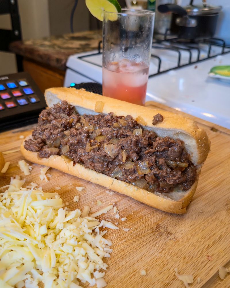 Homemade cheese steak with onions, cheese meat on a long hoagy, roll with cheese in the foreground and a refreshing drink in the background