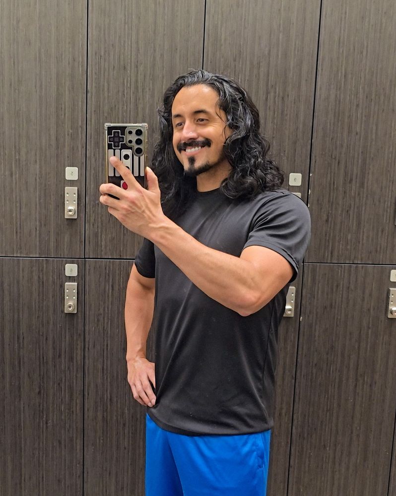 A man with long wavy, black hair is showing off athletic build in a gym amir selfie smiling because he's happy for healthy motivation