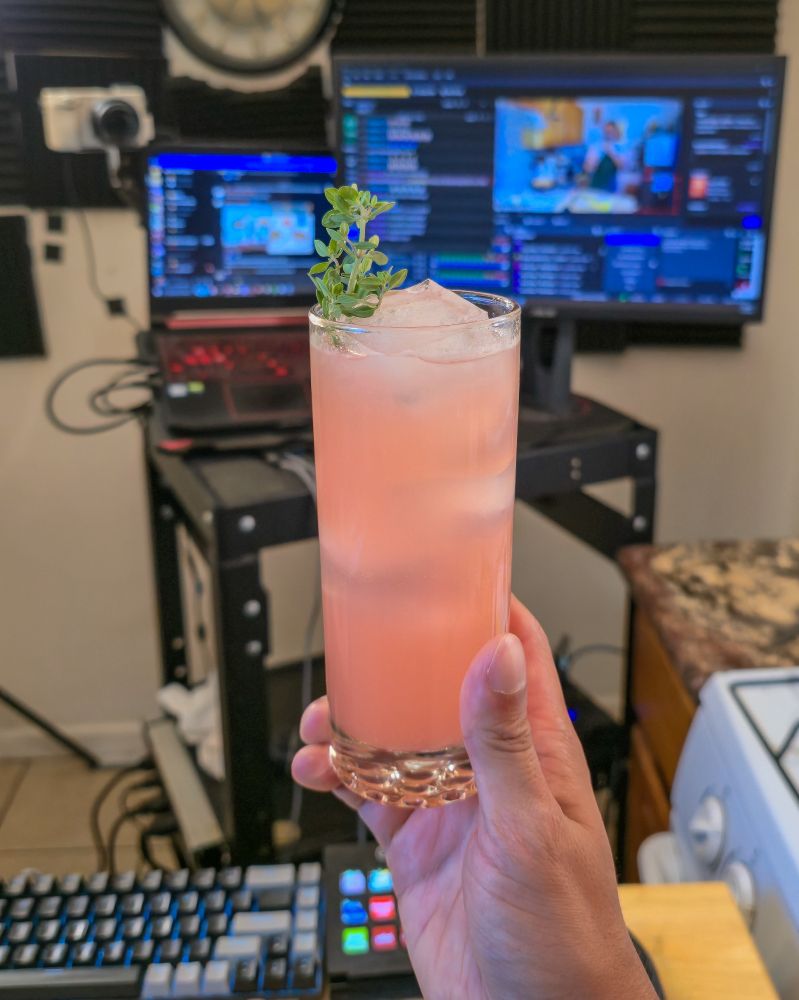 A peach and pink colored beverage, cold and with condensation and ice, with a twig of time sticking out of it in front of a keyboard, a stream deck and monitors that are blurry behind it, denoting, that it has been made on a twitch stream and being held up by a chef