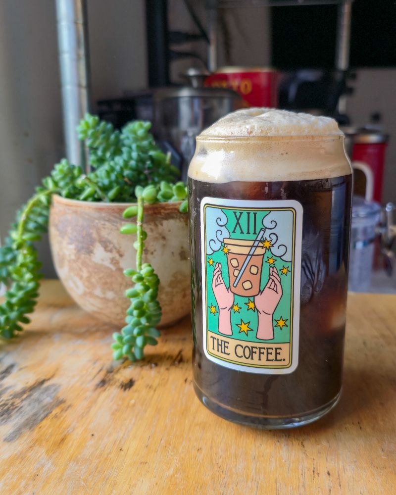 A frothy and cold iced coffee. It's in a glass mug that depicts a tarot card that says the coffee on it with a clay pot and a succulent plant inside