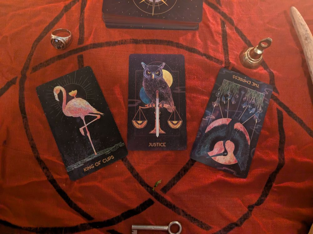 Three card tarot spread (King of Cups upright, Justice upright, & the Empress inverted) on an alter of red cloth with a triquetra print on it along with a ring, bell, key, stave, feather, sage smudge in a spider dish, and the rest of the tarot deck.