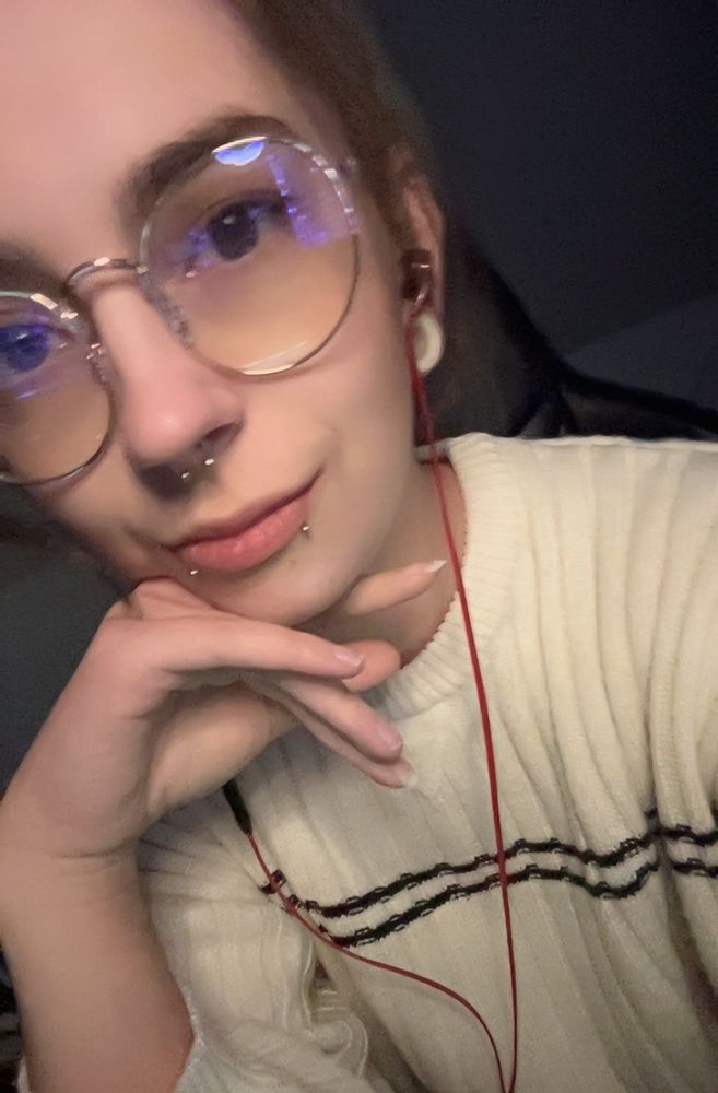 Selfie of me in a white sweater. I’m wearing gold rimmed glasses. My skin is white, my hair and eyes are brown. I have snakebite and septum piercings and have white tunnels in my ears.