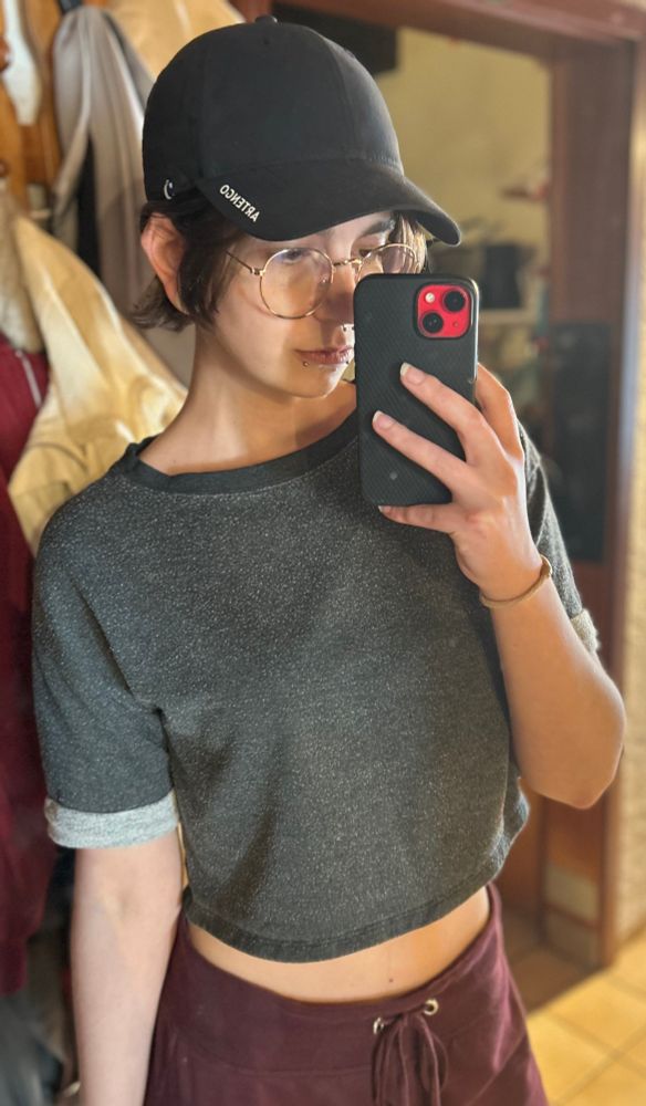 Mirror selfie of me in winered sweats and a black cropped shirt. I’m holding the phone slightly in front of my face. My skin is white, my eyes and hair are brown. I’m wearing gold rimmed glasses and have snakebite piercings and a septum piercing and white tunnels in my ears.