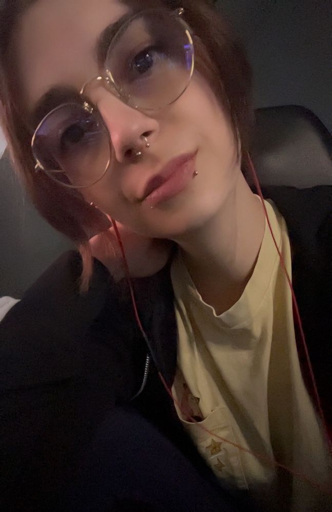 Selfie of me in a pastel yellow shirt and black hoodie whilst smiling. My skin is white, my hair and eyes are brown. I’m wearing gold rimmed glasses and have snakebite and septum piercings. I have white tunnels in my ears.