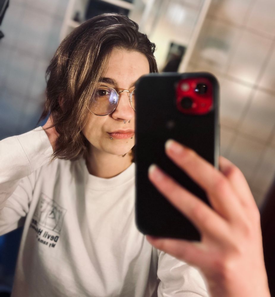 Mirror selfie of me in a white sweater. I’m wearing gold rimmed glasses. My skin is white, my hair and eyes are brown. I have snakebite and septum piercings and have white tunnels in my ears. I’m holding my phone in a black case in front of my face.