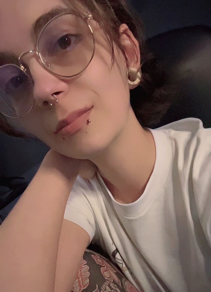 Selfie of me in a white shirt. I’m wearing gold rimmed glasses. My skin is white, my hair and eyes are brown. I have snakebite and septum piercings and have white tunnels in my ears.
