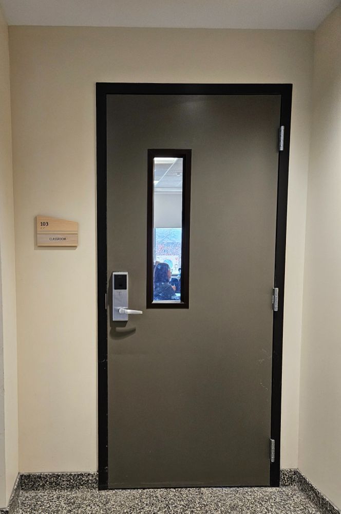 Closed gray door of a classroom. The door has a vertical window, and a person is visible inside the room.